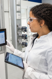 A woman in a laboratory coat and safety glasses interacts with a touchscreen display while holding a tablet, surrounded by equipment.