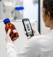 A person in a lab coat examines a brown bottle with a blue cap while using a smartphone to check information on the screen.