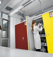 Eine Person in einem Labor mit einem weißen Kittel, die eine Chemikalie aus einem gelben Schrank holt. Im Hintergrund sind weitere Schränke und ein Tisch zu sehen.