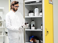 A scientist in a lab coat and safety goggles examines a bottle while standing in front of a cabinet filled with various chemical containers.