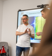 Zwei Personen stehen vor einem Präsentationsbildschirm. Eine Person hält ein Objekt in der Hand, während die andere etwas erklärt.