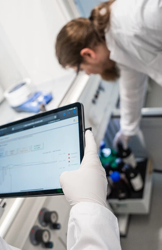 A laboratory scene featuring a person in a white lab coat and gloves holding a tablet displaying data, while another person is retrieving bottles from a nearby cabinet.