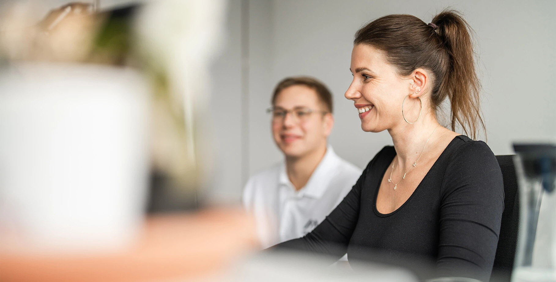 Two people are engaged in a conversation at a desk. One person, a woman with a ponytail and hoop earrings, is smiling while looking at a computer. The other person, a man with glasses, is smiling in the background. The setting appears to be an office environment.