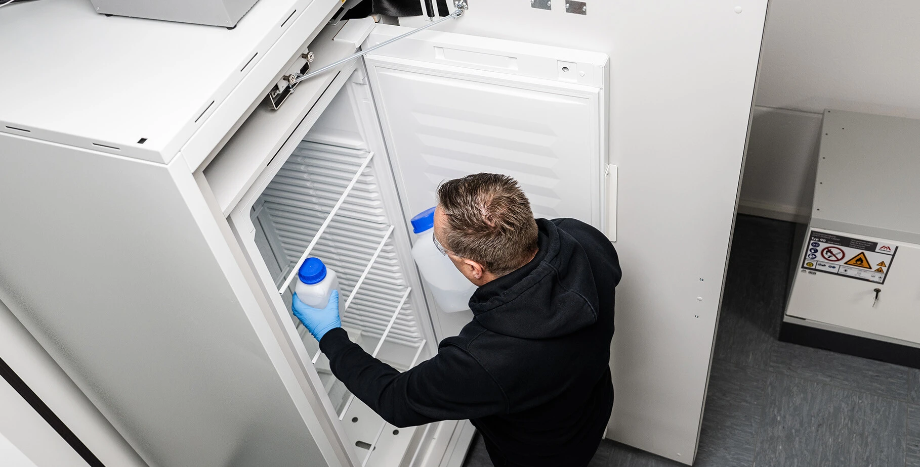 Mann in schwarzem Hoodie mit Schutzbrille, der eine Flasche in einen Gefrierschrank stellt.