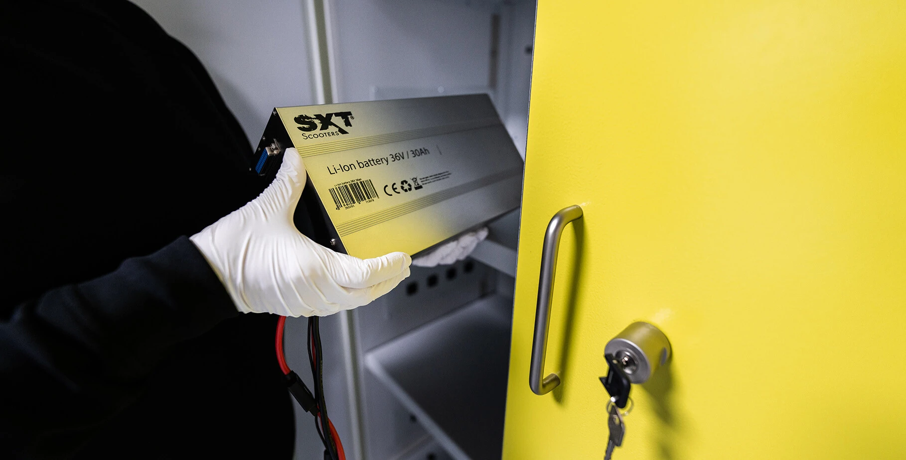 Ein Mensch in weißen Handschuhen hält eine Li-Ion-Batterie und platziert sie in einen gelben Schrank.
