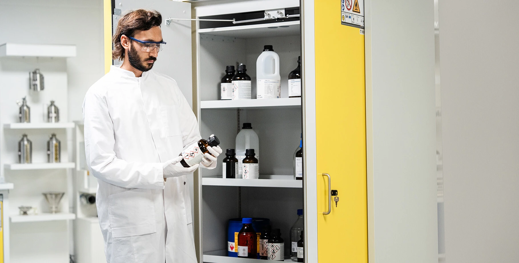 Ein Laborant in einem weißen Kittel steht vor einem Schrank voller Chemikalienflaschen und hält eine Gefahrstoffflasche in der Hand.