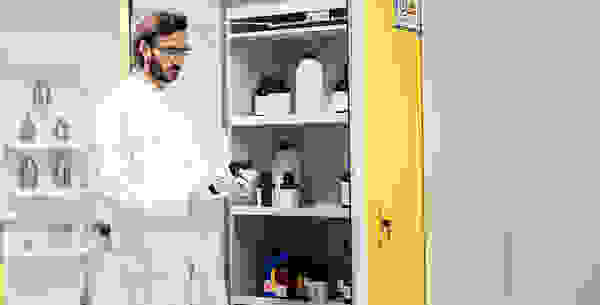 A man in a lab coat and safety goggles is holding a bottle while examining shelves filled with various chemical containers in a laboratory setting.