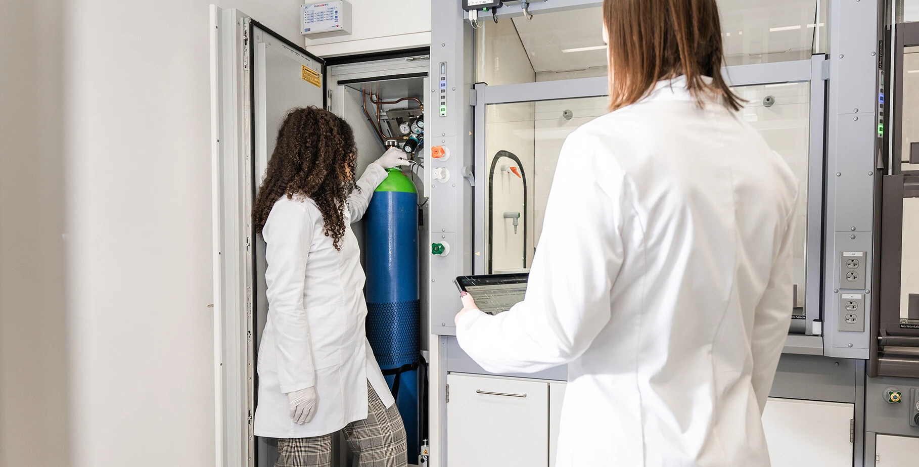 Two lab-coated individuals are working with a gas cylinder inside a lab environment, one handling the cylinder and the other holding a tablet.