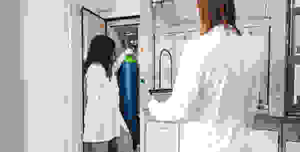 Zwei Personen in Laborkitteln arbeiten in einem Labor. Eine Person befindet sich vor einem Gerät und handhabt einen Farbkessel, während die andere Person ein Tablet hält.