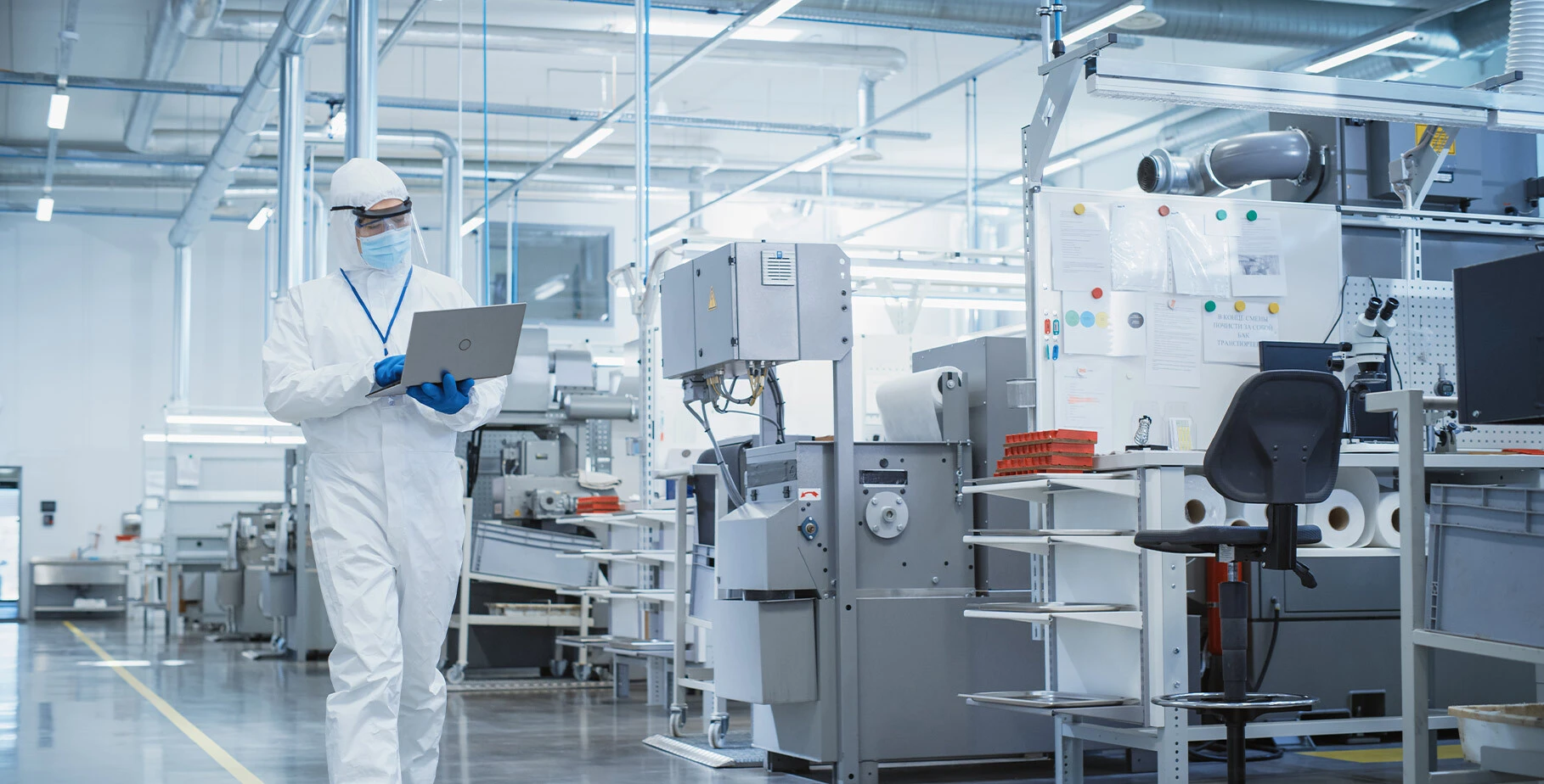 A person in a white lab coat and protective gear is using a laptop in a cleanroom environment filled with equipment and workstations.