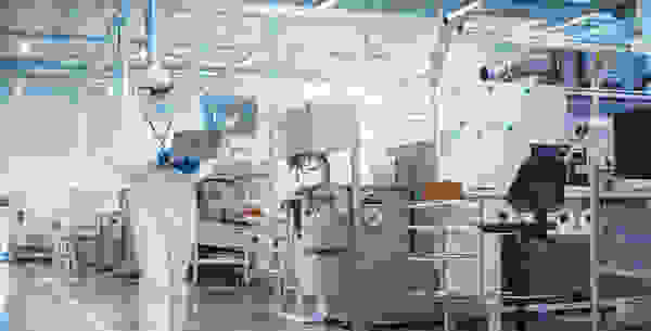 A person in a white lab coat and protective gear is using a laptop in a cleanroom environment filled with equipment and workstations.