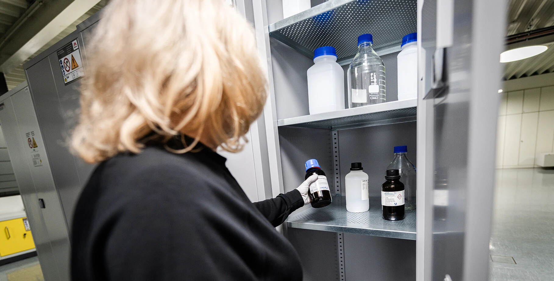A person wearing gloves is inspecting a shelf filled with various labeled laboratory bottles in a storage cabinet.