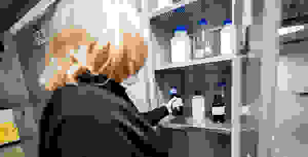 A person wearing gloves is inspecting a shelf filled with various labeled laboratory bottles in a storage cabinet.