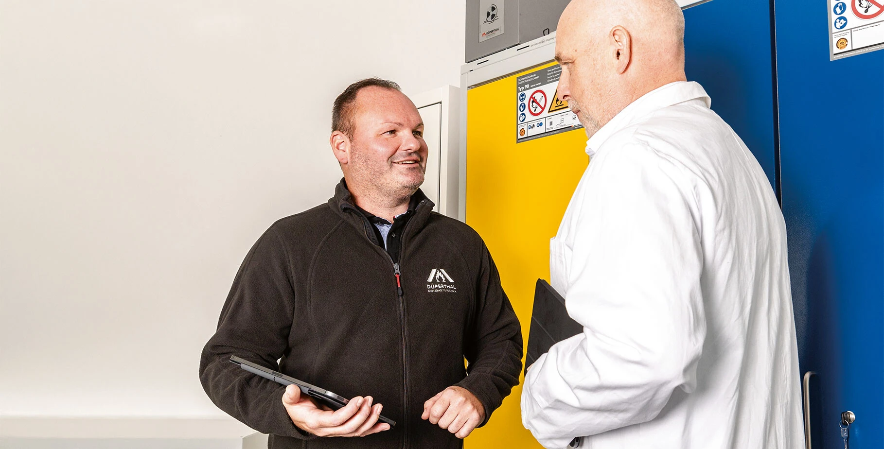 Two men are engaged in conversation in a professional setting, with one holding a tablet. Behind them are brightly colored cabinets, one yellow and one blue.