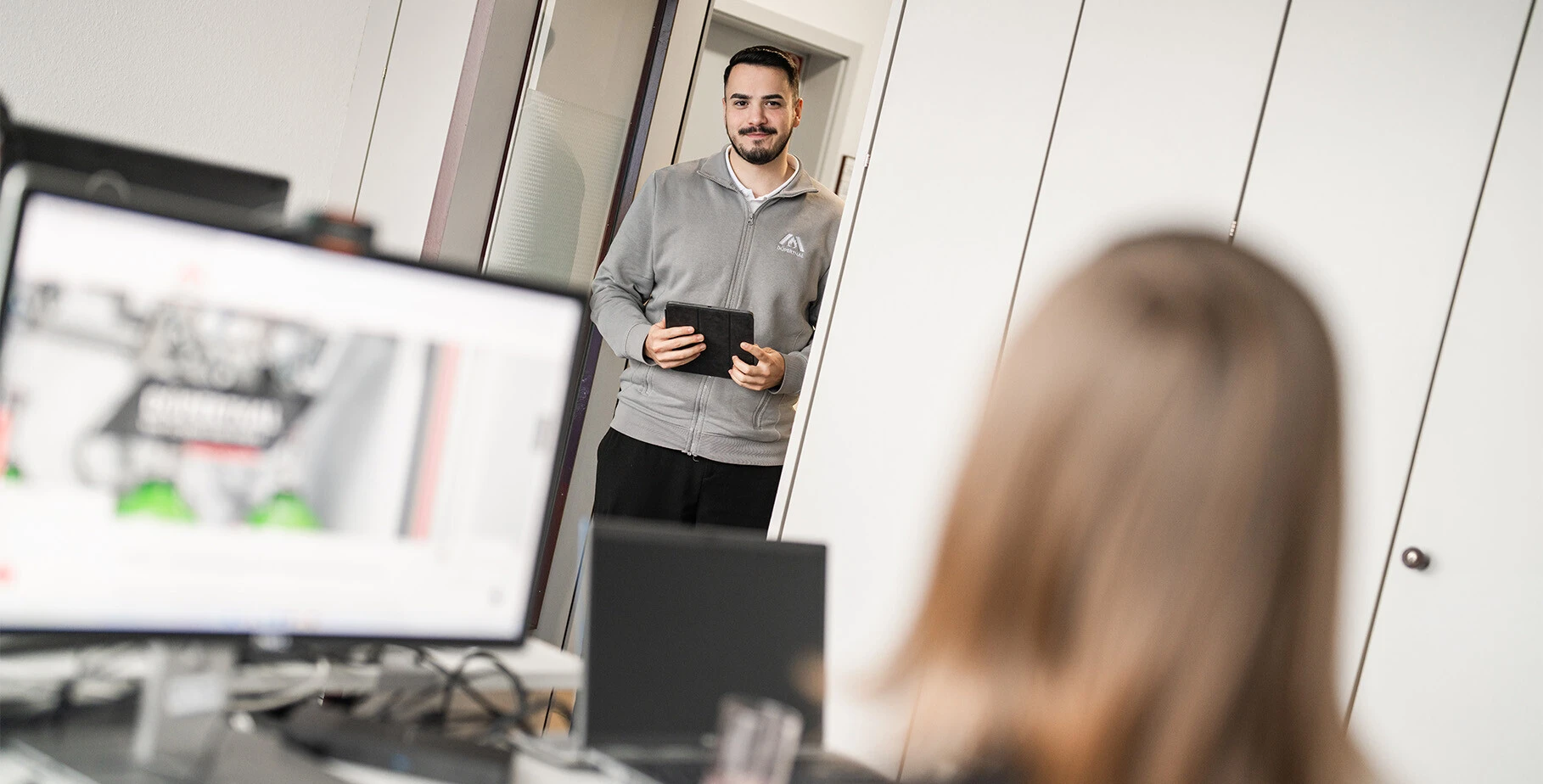 Ein Mann steht in einem Büroraum und hält ein Tablet in der Hand, während ein Schreibtisch mit Computerbildschirmen im Vordergrund zu sehen ist