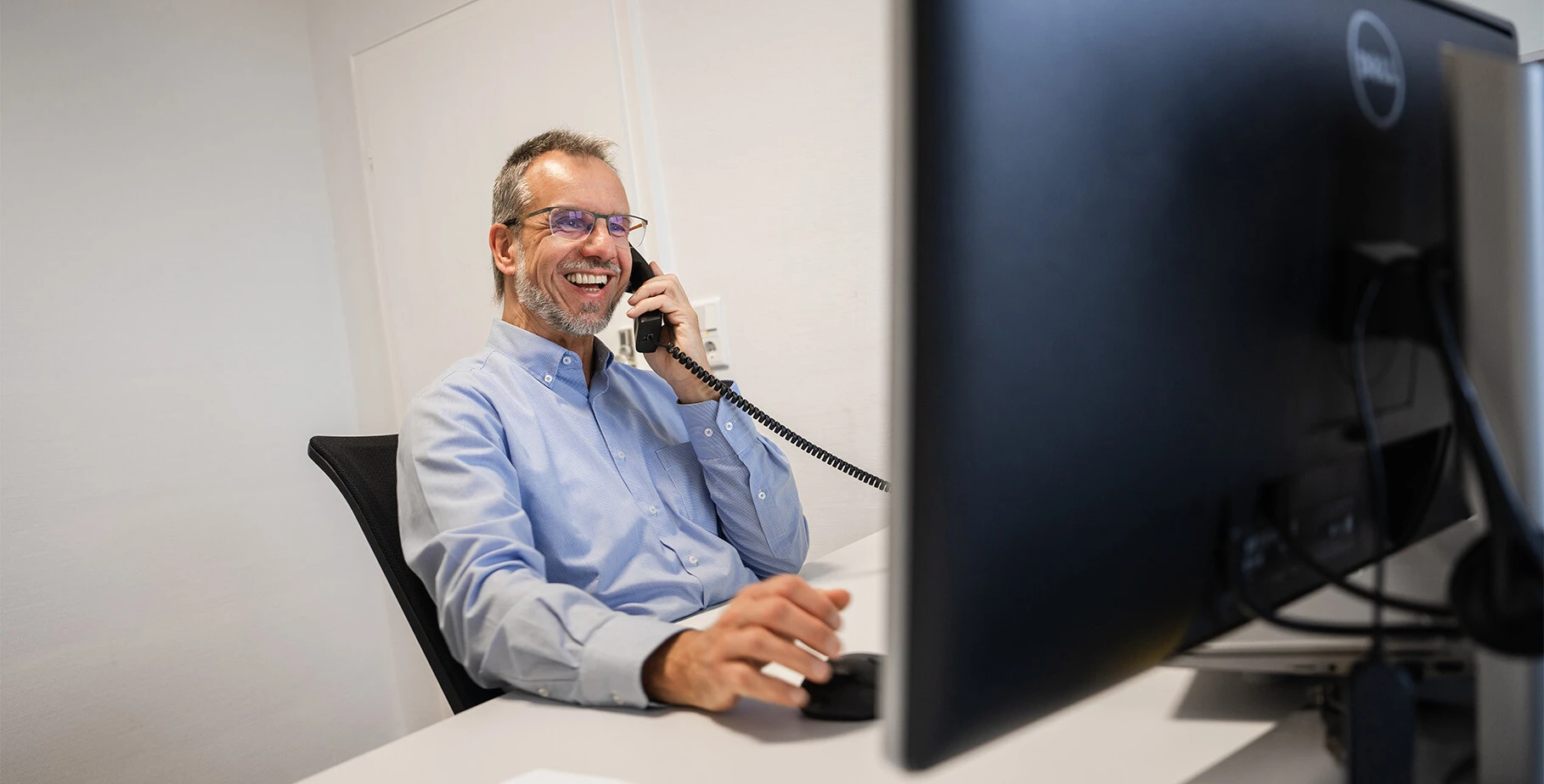 Ein lächelnder Mann im Büro spricht am Telefon und arbeitet an einem Computer.