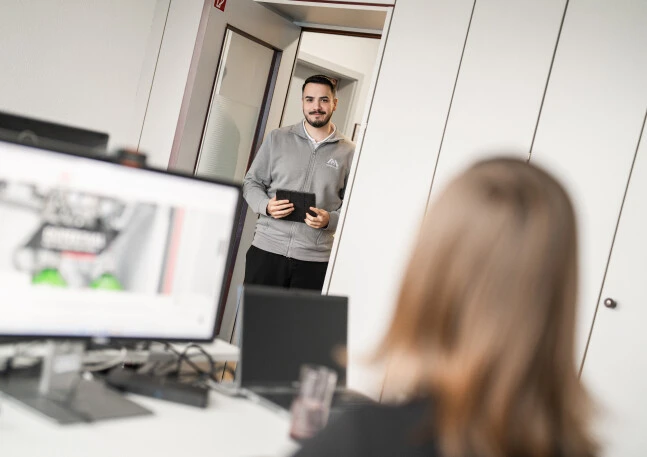 A man stands in a doorway holding a tablet, while another person with long hair looks away from him. Computer screens can be seen in the foreground.