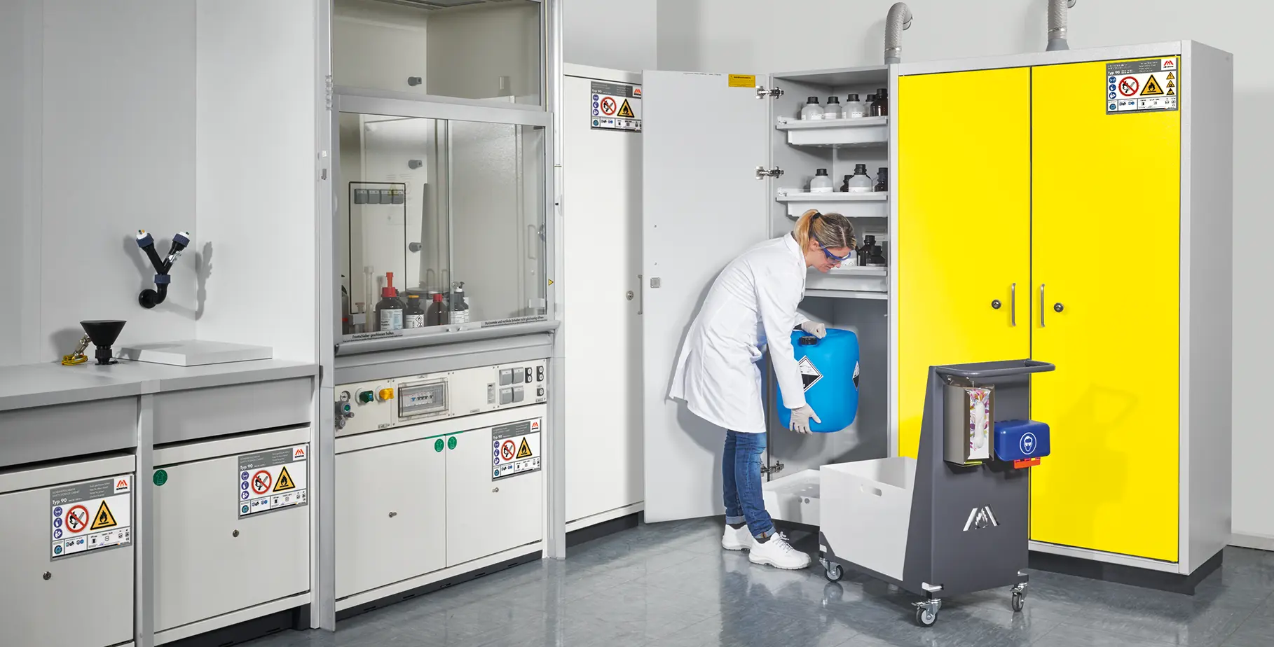 Laboratory mit einer Person in einem weißen Laborkittel, die einen blauen Behälter in einen Schrank mit Warnhinweisen stellt. Im Hintergrund sind weitere Labormöbel und Ausstattungen sichtbar.