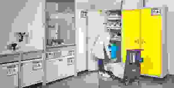 A laboratory setting featuring a lab technician in a white coat and safety goggles, storing a blue container in a cabinet with a yellow door. Surrounding equipment includes a fume hood, various storage cabinets, and safety labels.