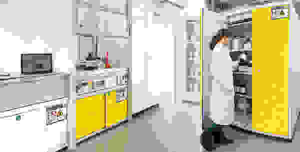 Laborszene mit einer Person in einem weißen Kittel, die Chemikalien aus einem gelben Schrank entnimmt. Hintergrund zeigt Laborausrüstung und einen Laptop.