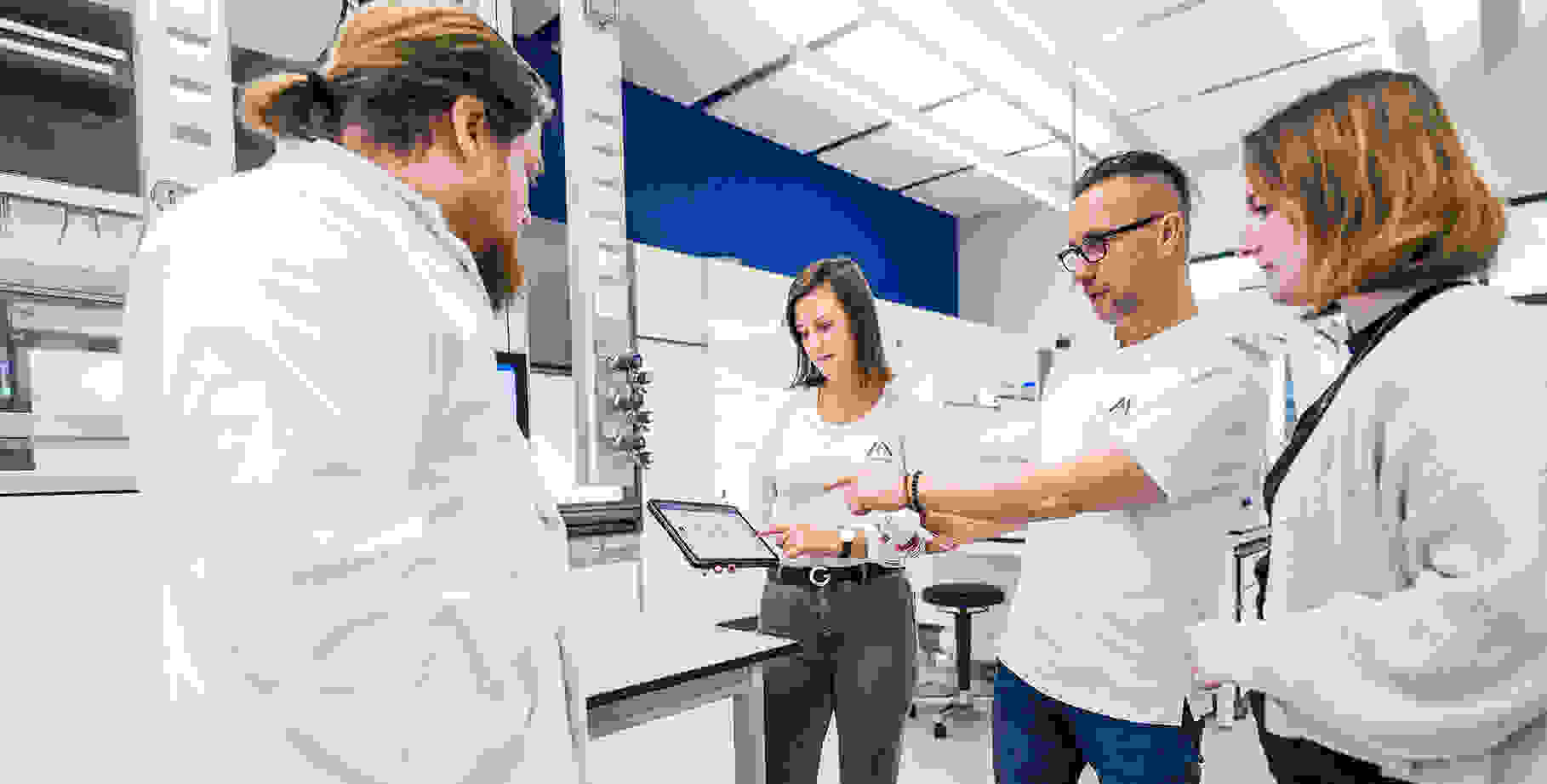 A group of four individuals in lab coats is discussing data displayed on a tablet in a modern laboratory setting.