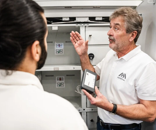 Zwei Männer diskutieren über ein Gerät in einem technischen Raum. Der ältere Mann zeigt mit einer Hand auf das Gerät, während er es in der anderen hält.