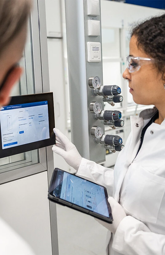 Zwei Personen in Laborkleidung verwenden ein Tablet und ein Display zur Überwachung von Geräteparametern in einem Labor.