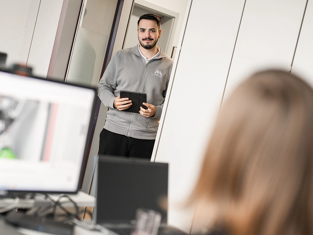 Ein Mann steht in einem Büroraum und hält ein Tablet in der Hand, während ein Schreibtisch mit Computerbildschirmen im Vordergrund zu sehen ist
