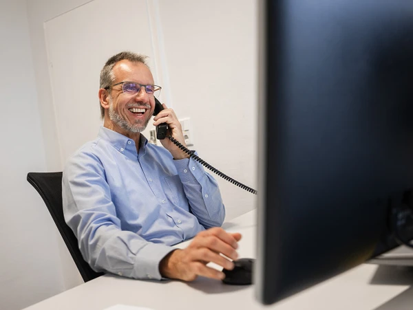 Ein lächelnder Mann im Büro spricht am Telefon und arbeitet an einem Computer.