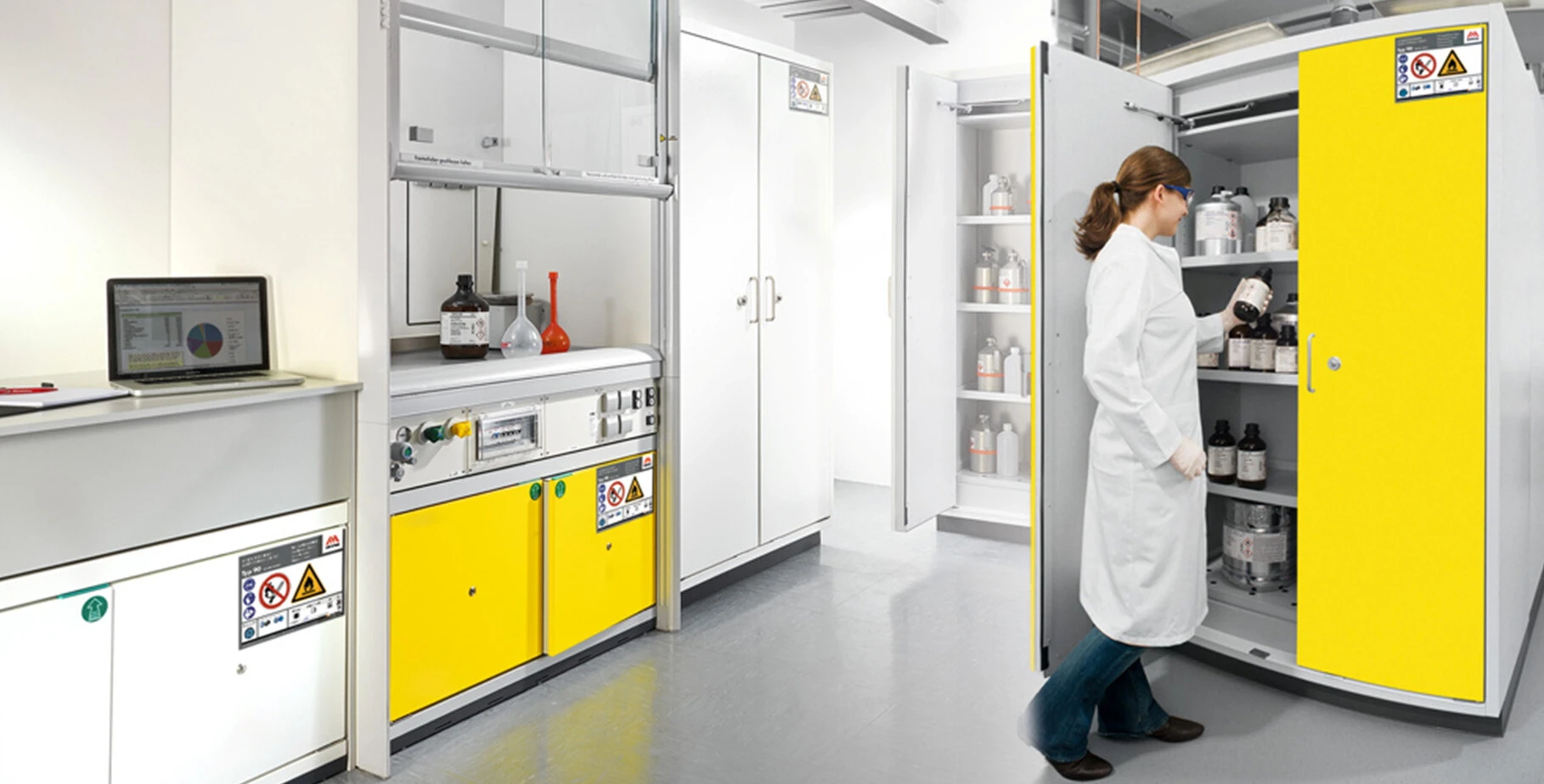 Laboratory scene with a person in a white coat taking chemicals from a yellow cabinet. The background shows laboratory equipment and a laptop.