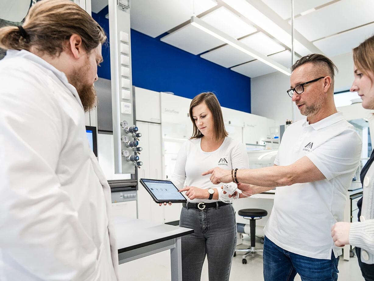 A group of four individuals in lab coats is discussing data displayed on a tablet in a modern laboratory setting.