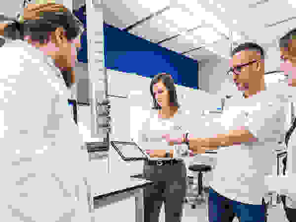 A group of four individuals in lab coats is discussing data displayed on a tablet in a modern laboratory setting.