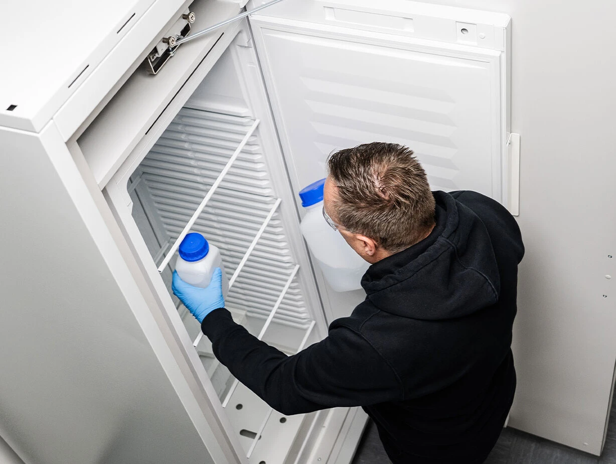 Ein Mann mit Handschuhen hält einen Behälter in einer leeren Kühlschranktür.