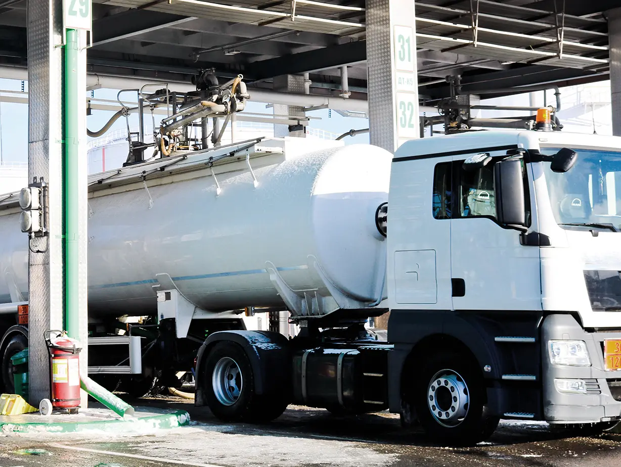 Ein Lkw mit einem großen Tank auf einer Tankstelle, umgeben von Rohrleitungen und Pumpen.