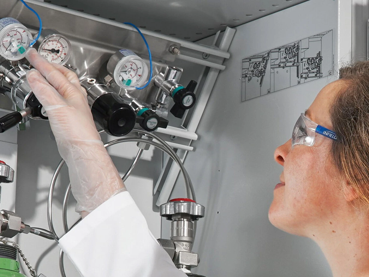 Eine Person in Schutzbrille und Handschuhe bedient ein Gasregelsystem mit Druckanzeigen und Ventilen.