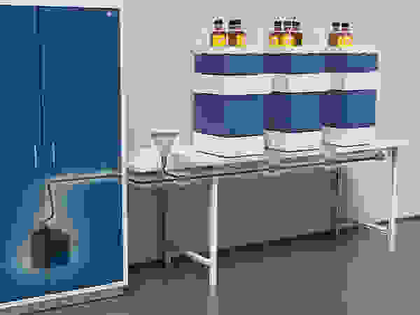 A blue storage cabinet is shown alongside a white table with stacked containers. Some containers have hoses attached, and a funnel is positioned on the table, indicating a setup for liquid transfer or chemical storage.