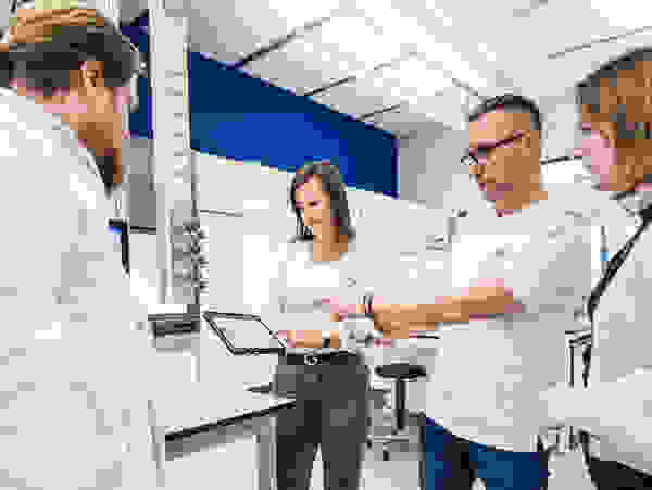 Vier Personen in einem Labor schauen auf ein Tablet. Sie diskutieren, während Geräte und Laborausrüstung im Hintergrund zu sehen sind