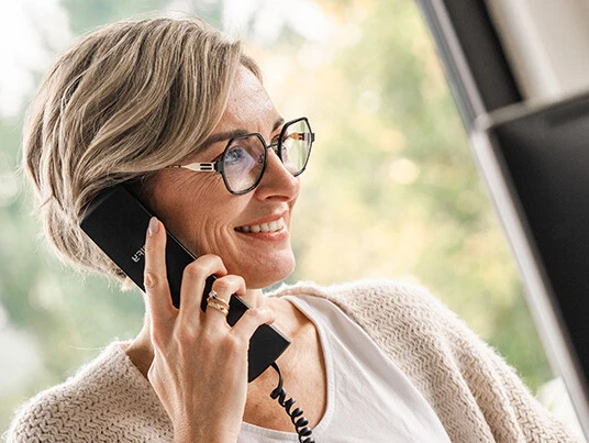 Frau mit kurzen Haaren und Brille, die lächelnd telefoniert, trägt einen hellen Cardigan.