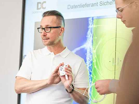 Zwei Männer präsentieren ein Gerät in einem Konferenzraum, während im Hintergrund ein Bildschirm mit Diagrammen und Informationen zu sehen ist.