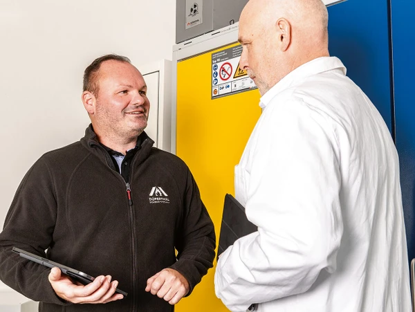 Two men are engaged in conversation in a professional setting, with one holding a tablet. Behind them are brightly colored cabinets, one yellow and one blue.