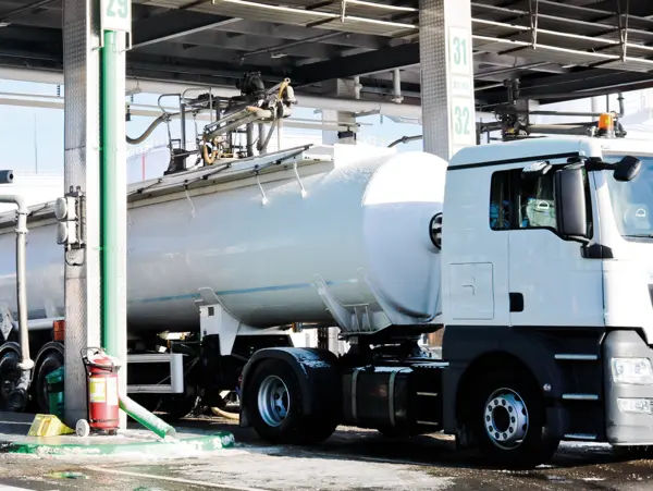 A white tanker truck is refueling at a gas station, with hoses and equipment for dispensing fuel visible.