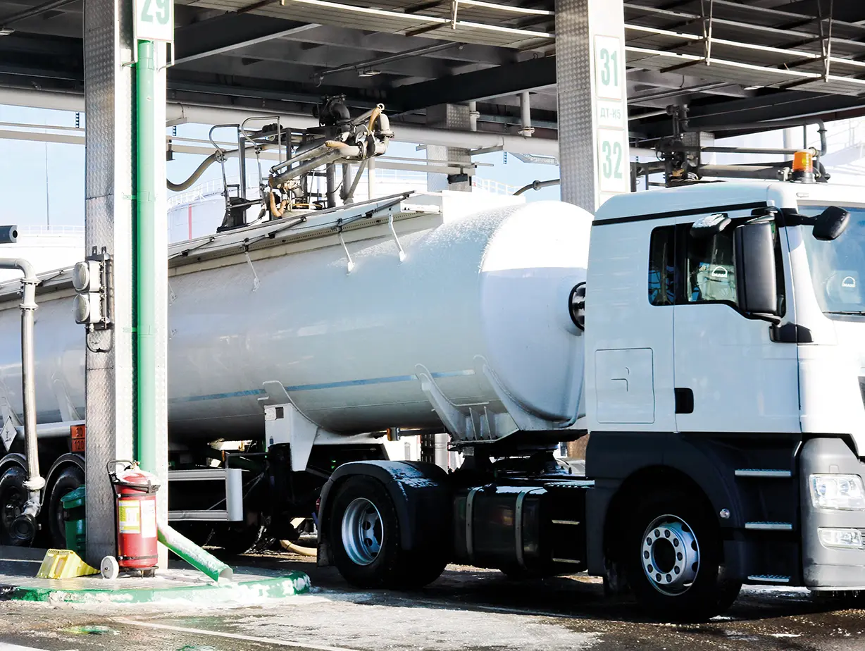A white tanker truck is refueling at a gas station, with hoses and equipment for dispensing fuel visible.