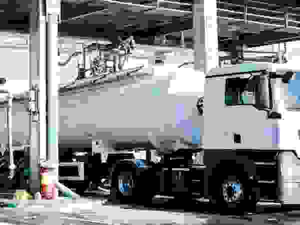 A white tanker truck is refueling at a gas station, with hoses and equipment for dispensing fuel visible.