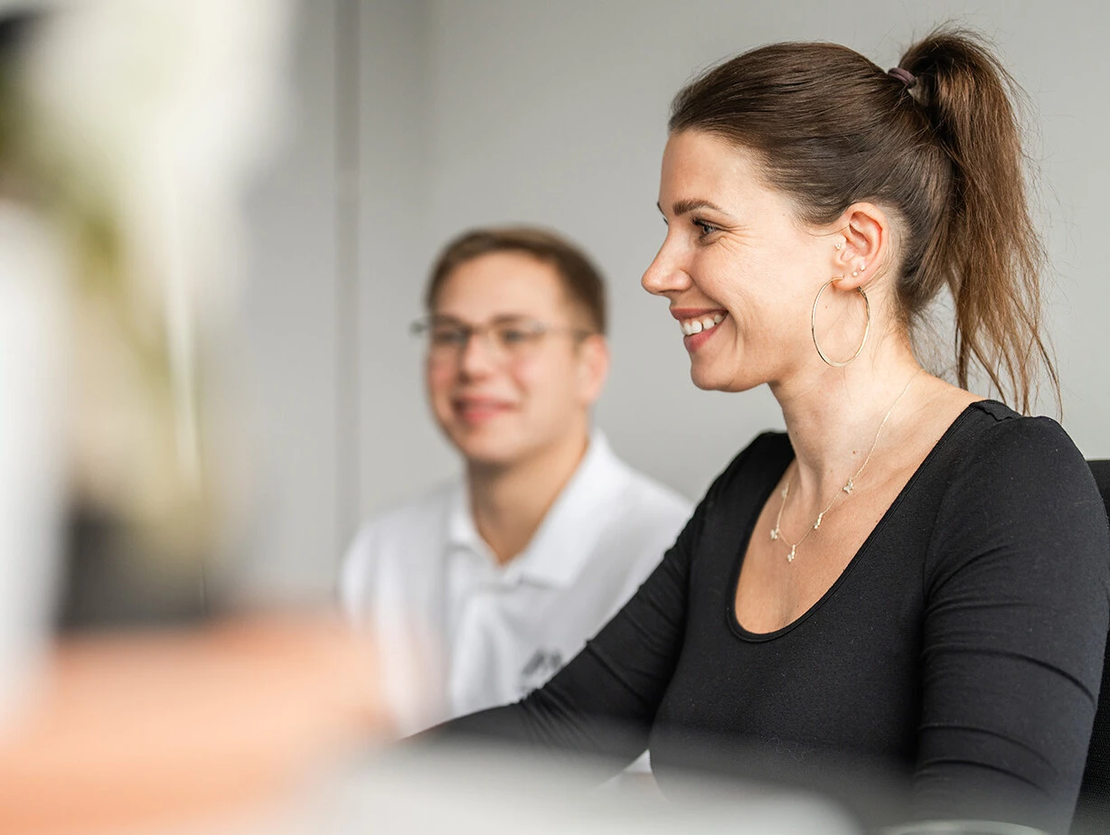 Two people are engaged in a conversation at a desk. One person, a woman with a ponytail and hoop earrings, is smiling while looking at a computer. The other person, a man with glasses, is smiling in the background. The setting appears to be an office environment.
