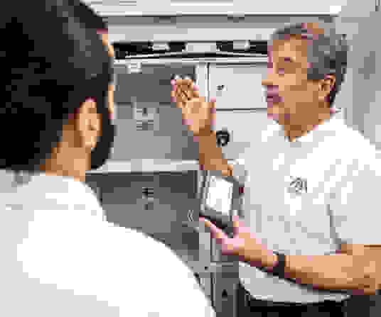 A man in a white polo shirt is explaining something while holding a device, with a cabinet in the background. Another person, mostly out of focus, is listening attentively.