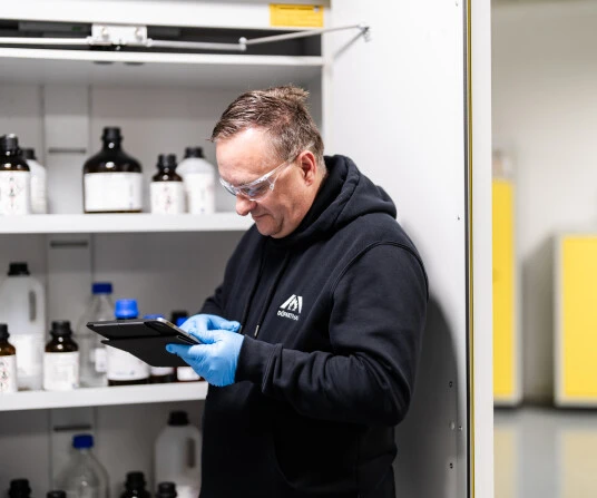 Ein Mann mit Schutzbrille und Handschuhen steht neben einem Regal mit Chemikalien und schaut auf ein Tablet.