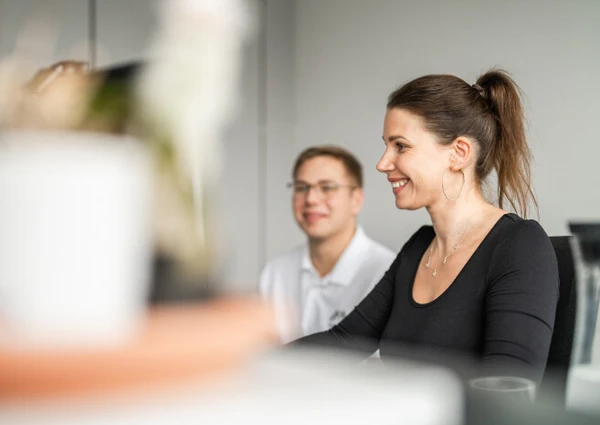 Ein lächelndes Paar sitzt in einem modernen Büro, der Fokus liegt auf der Frau mit langen Haaren und OHrringen, die am Computer arbeitet. Der Mann im Hintergrund lächelt ebenfalls.