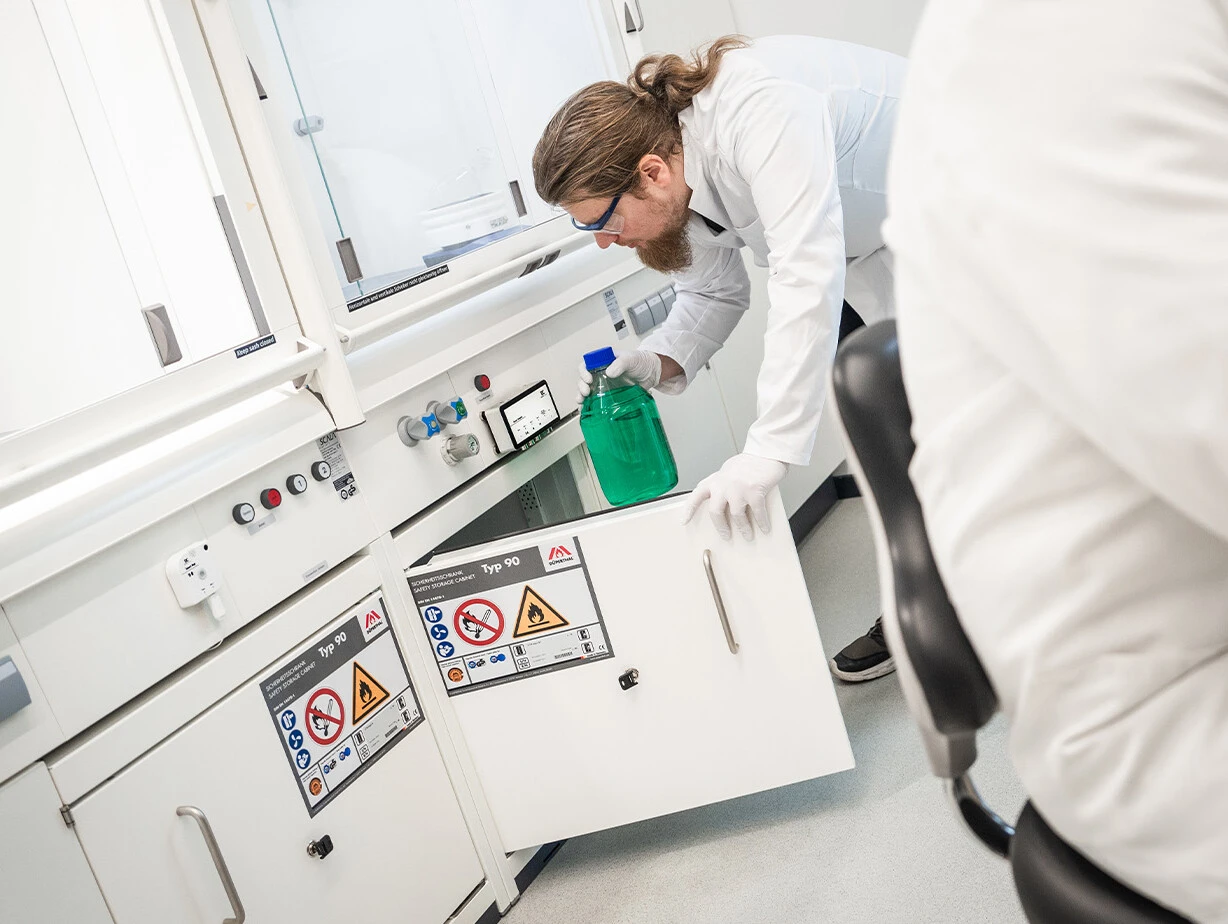 Eine Person im Laborkittel und mit Handschuhen greift nach einer grünen Flüssigkeit in einem Labor. Im Hintergrund sind Laboreinrichtungen und Sicherheitszeichen sichtbar.