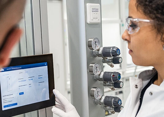 Zwei Personen in Laborkleidung verwenden ein Tablet und ein Display zur Überwachung von Geräteparametern in einem Labor.
