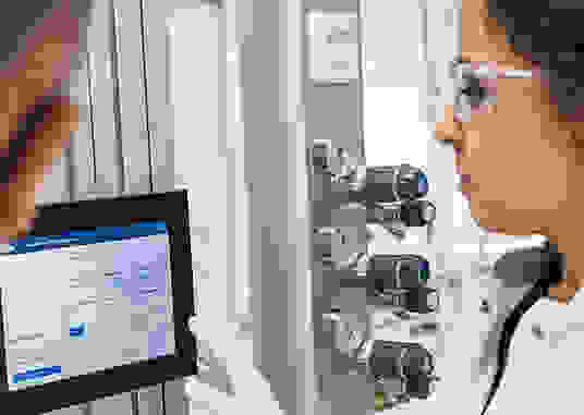 A laboratory setting featuring a person in a white coat using a tablet and interacting with a touchscreen display, alongside pressure gauges on a panel.